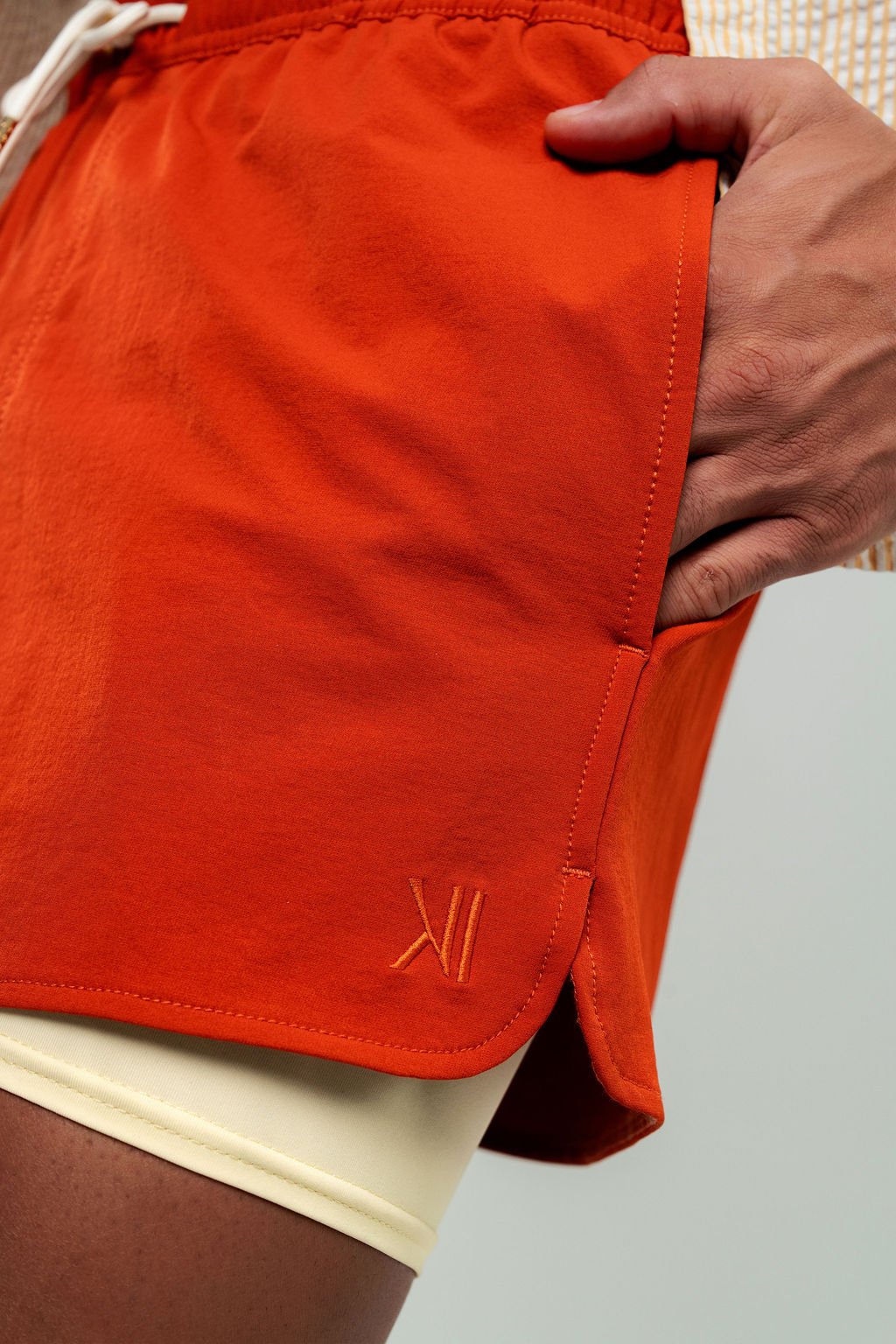 Close-up of DUSKITTEN logo embroidery on the right hem of ECLIPSE LINE swim shorts in burnt orange, showing side pocket construction and buttery yellow inner lycra type liner, Duskened Genesis collection.